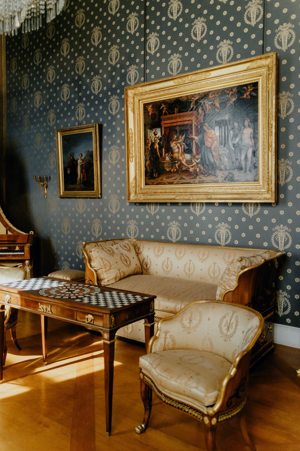 Living room with blue and gold ornate damask wallpaper, an antique cream sofa and gilt-framed art